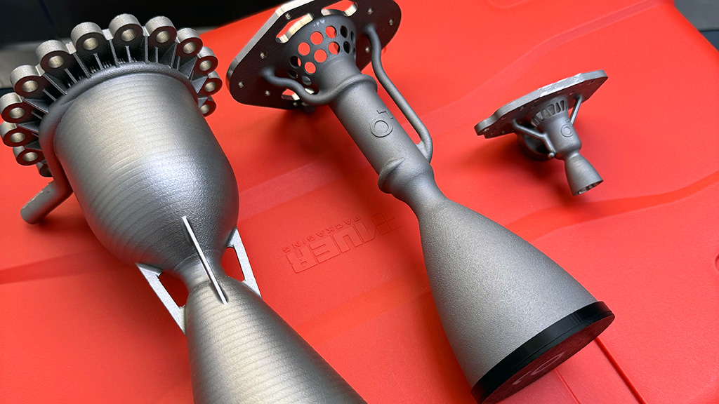 Three gray, metallic, 3D-printed rocket engine nozzles, varying in size, are displayed on a bright red surface. The largest nozzle on the left has a wide top with multiple circular ports. The middle nozzle is taller and more slender, featuring a flared base and a complex, branched manifold at its top. The smallest nozzle on the right is similar in design to the middle one but proportionally smaller. All three components exhibit a textured metallic finish, indicative of additive manufacturing. They are produced by Liftero.